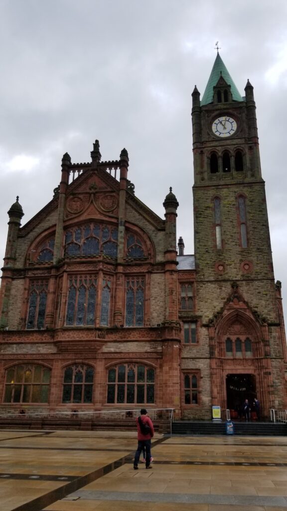 Overcast day view of the historic Guildhall in Derry, with its distinct red sandstone facade and neo-gothic architecture, featuring a lone individual in the foreground.