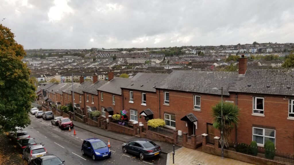 Overcast view of a residential street in Derry with a row of red brick houses, parked cars, and a backdrop of densely packed houses extending into the distance.