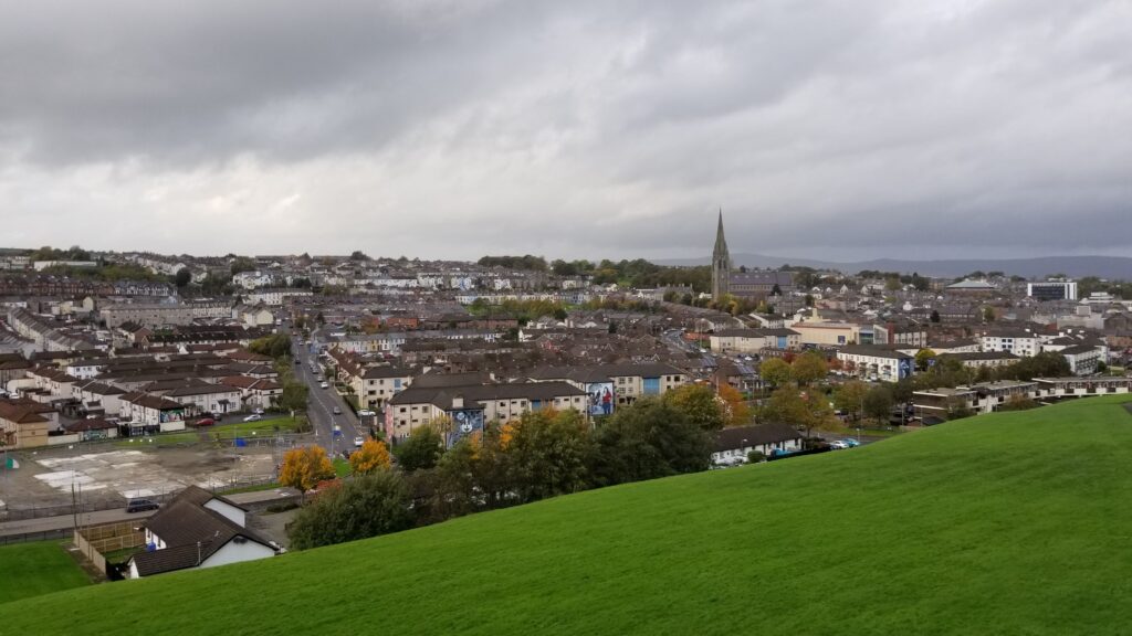 Panoramic view of Derry-Londonderry, showing the dense urban landscape with rows of houses and the prominent spire of St. Eugene's Cathedral in the distance under a cloudy sky.