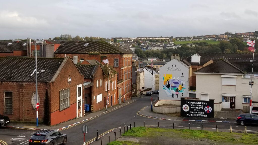 A view of a street in Derry with historical murals, including one featuring the slogan 'LondonDerry West Bank Loyalists Still Under Siege No Surrender,' depicting the city's complex political history.