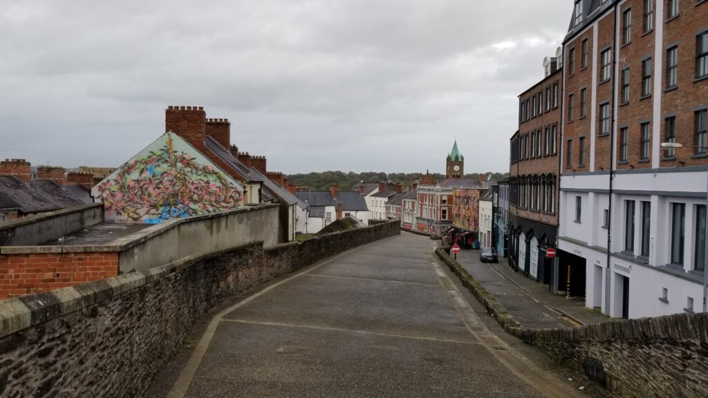 A street-level view of Derry's walls with a vibrant street art mural, traditional terraced houses, and the Guildhall's clock tower in the background, under a grey sky.