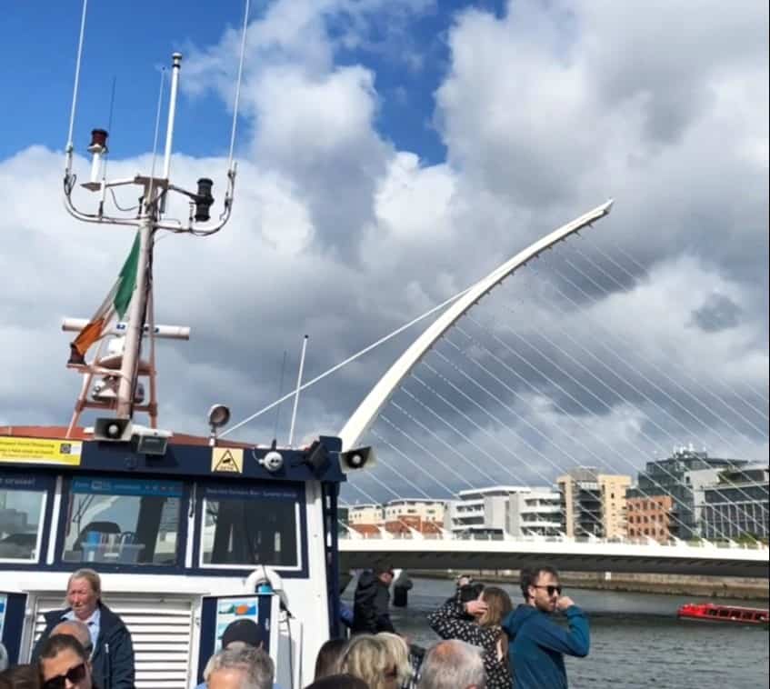 Passengers aboard a Dublin Bay cruise boat, with the iconic Samuel Beckett Bridge in the background. The Irish flag flutters atop the boat's mast, set against a backdrop of partly cloudy skies, while a photographer captures the moment, illustrating a popular tourist activity in Dublin.