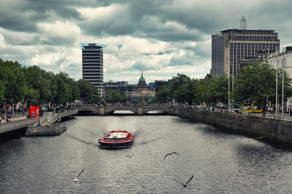 A red sightseeing boat cruises along the River Liffey in Dublin, passing under an arched stone bridge. The city's dynamic architecture, including a distinctive round-topped building and modern skyscrapers, frames the riverbanks, while several seagulls glide over the water, adding life to this urban scene.