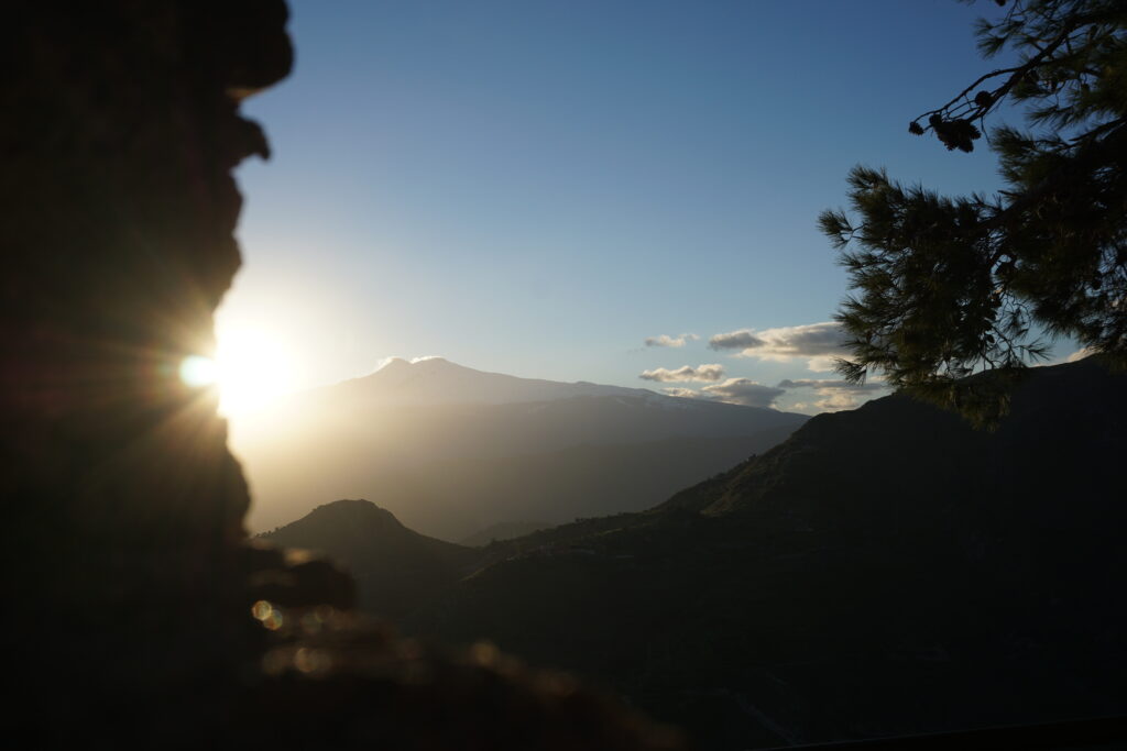 The sun setting behind Mount Etna, casting a soft glow and creating a silhouette of the mountain against a hazy sky, with the foreground featuring the dark outline of a rocky edge and pine branches.