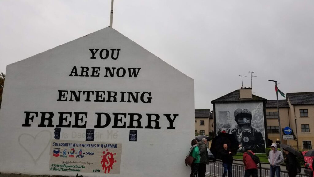 The iconic 'You Are Now Entering Free Derry' mural on a white gable wall, with people gathered around it, symbolizing the city's civil rights history and political murals.