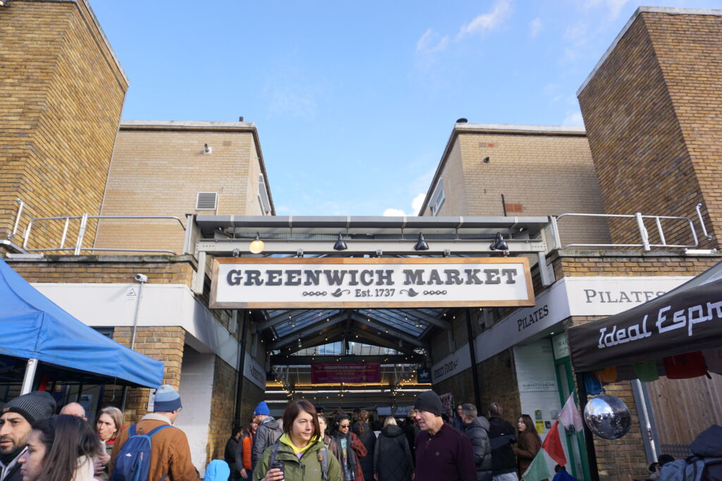 he entrance of Greenwich Market in London, bustling with visitors under a sign that reads 'Greenwich Market Est. 1737', set against a clear blue sky with surrounding brick buildings.