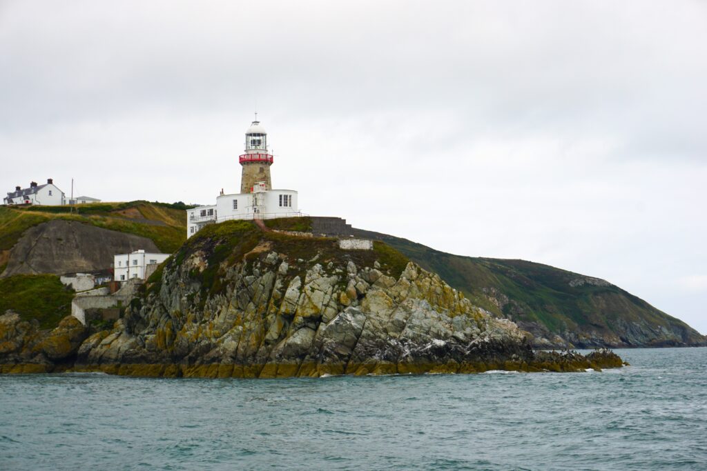 The Howth Lighthouse stands prominently on a grassy headland, overlooking the sea with its white and red structure providing a stark contrast to the overcast sky. Jagged rocks line the water's edge, and the calm sea reflects the grey tones of the sky, capturing the peaceful yet rugged coastal landscape.