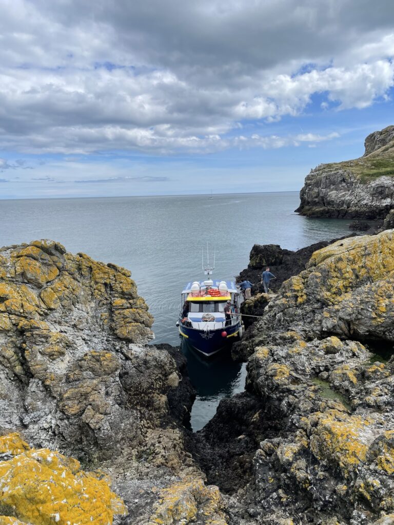 A tour boat is nestled between rugged, lichen-covered rocks at a docking area on Ireland's Eye, under a sky filled with soft clouds. The serene sea extends into the horizon, capturing the tranquil and adventurous essence of the island's boat tours.