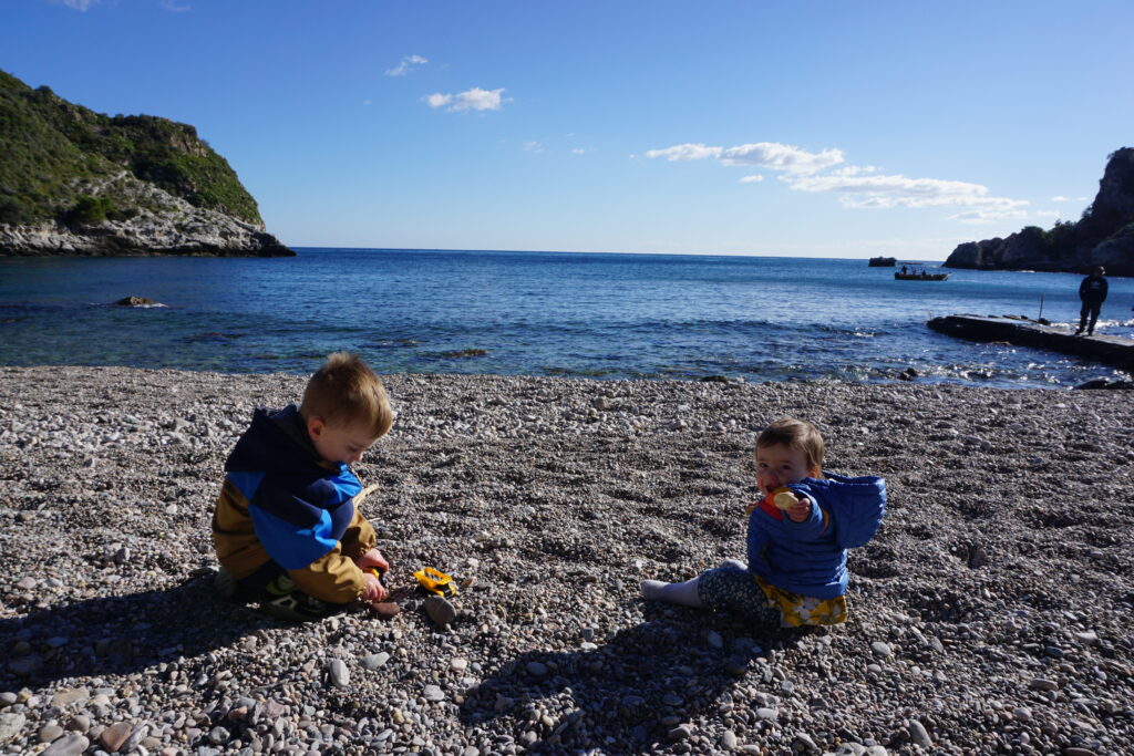 Two young children playing on a pebbled beach, one with a toy truck, against the backdrop of a serene sea and a rocky outcrop under a clear blue sky.