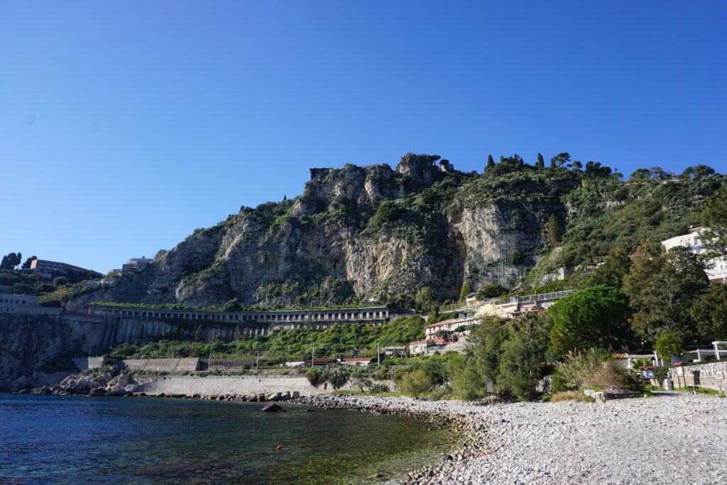 A panoramic view of Isola Bella in Taormina, showing the rugged cliffside adorned with greenery, a clear blue sea, and a pebbled beachfront.