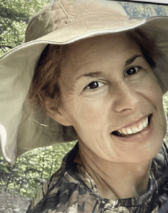 Close-up portrait of a smiling woman wearing a wide-brimmed hat, with a blurred greenery background, conveying a sense of adventure and warmth.