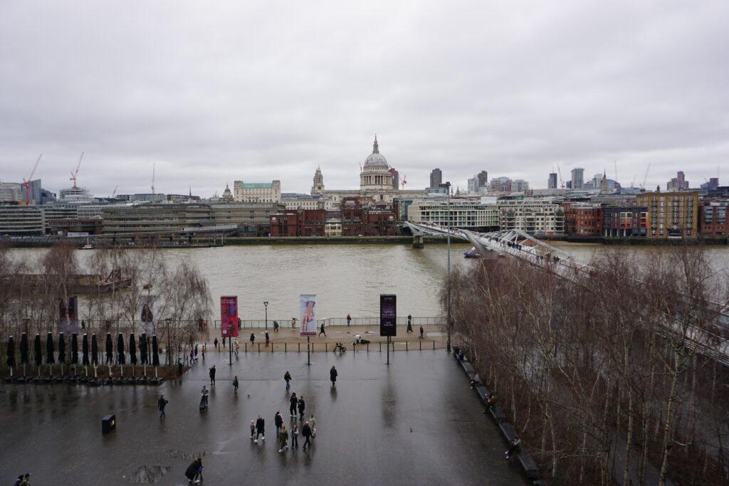 Overcast view from the South Bank overlooking the River Thames in London, with St. Paul's Cathedral in the background, the Millennium Bridge leading across the river, and people walking along the promenade.