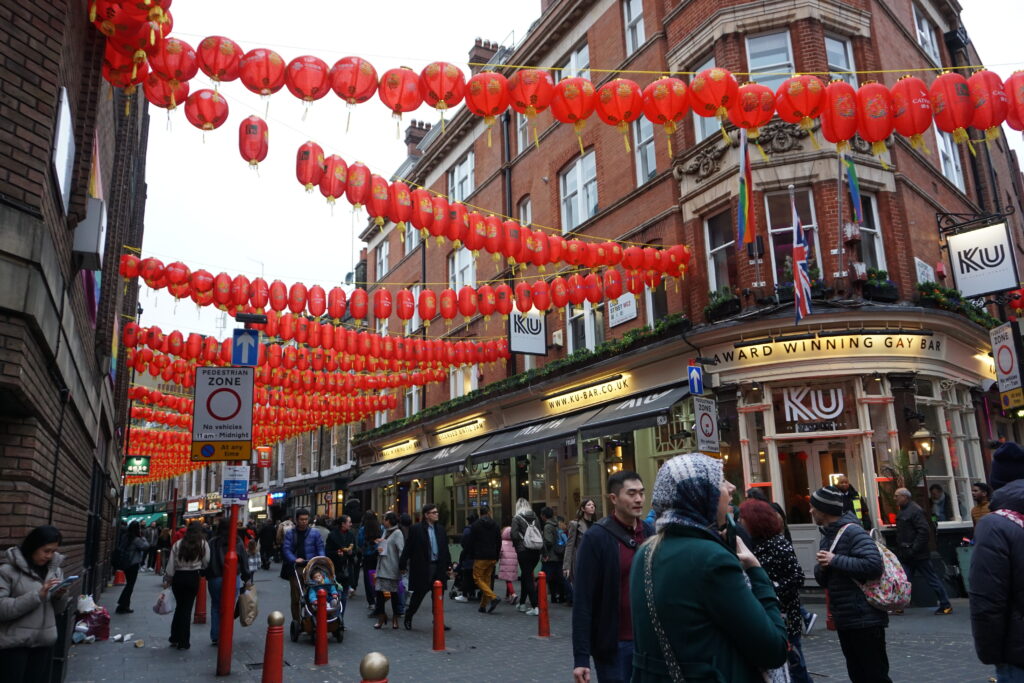 A vibrant scene from London's Chinatown decorated with red lanterns, with pedestrians walking through the pedestrian zone and a sign for an award-winning gay bar in the background.