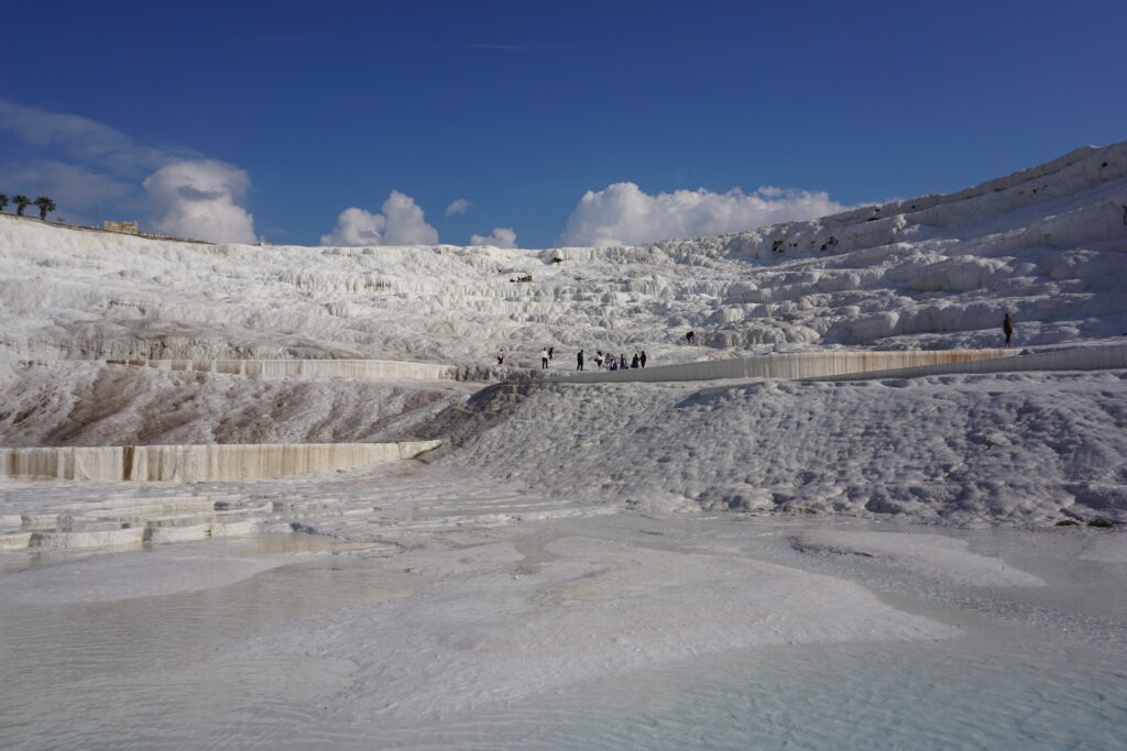 "Terraced mineral baths of Pamukkale in Turkey, with visitors walking along the white calcite-laden cliffs under a bright blue sky.