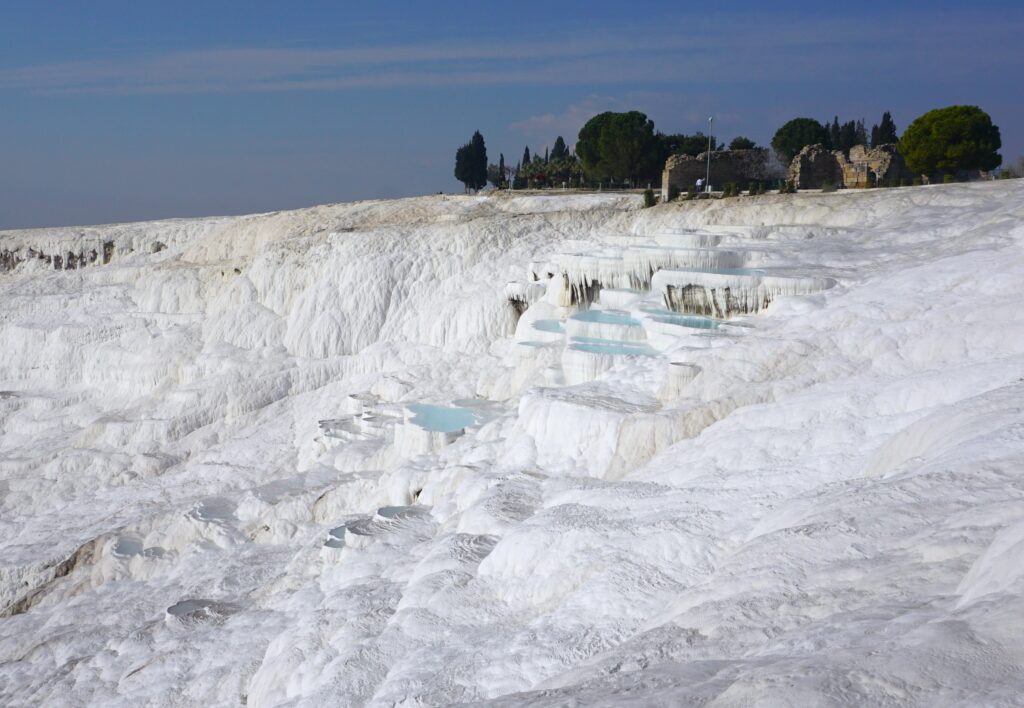 A stunning view of the pristine white terraces and thermal water pools of Pamukkale, Turkey, under a clear blue sky with historical ruins visible in the background atop the hillside.