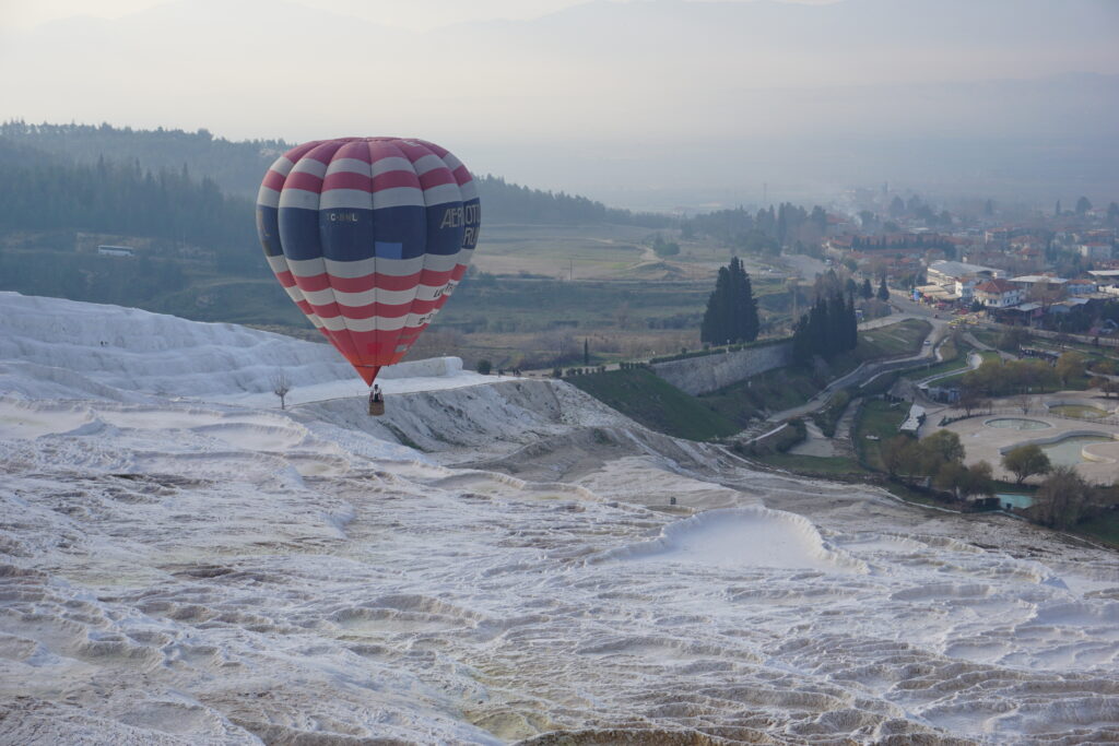 A hot air balloon featuring red, white, and blue stripes floats above the white terraces of Pamukkale, with a hazy backdrop of trees and a rural landscape.