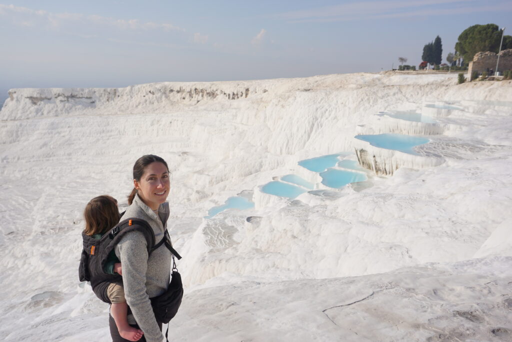 A woman, the author, smiling at the camera, carries a child on her back with a panoramic view of the Pamukkale travertine pools and terraces stretching behind them.