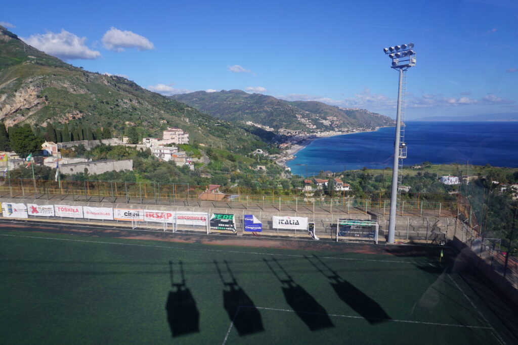 Aerial view from a cable car showing the shadows of the carriages on a green soccer field, with a backdrop of the Mediterranean Sea, a curving coastline, and the lush hills of Taormina.