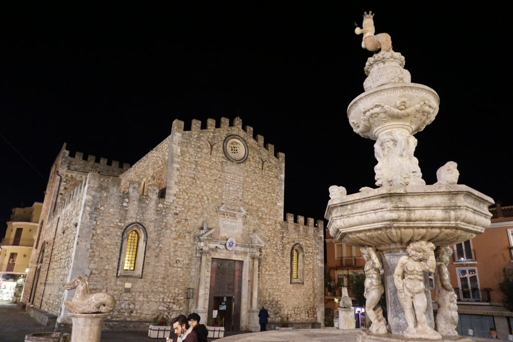 The historic Taormina Cathedral at night, illuminated by street lights, showcasing its medieval stone architecture and a detailed baroque fountain in the foreground with sculptures and an obelisk topped by a heraldic animal.