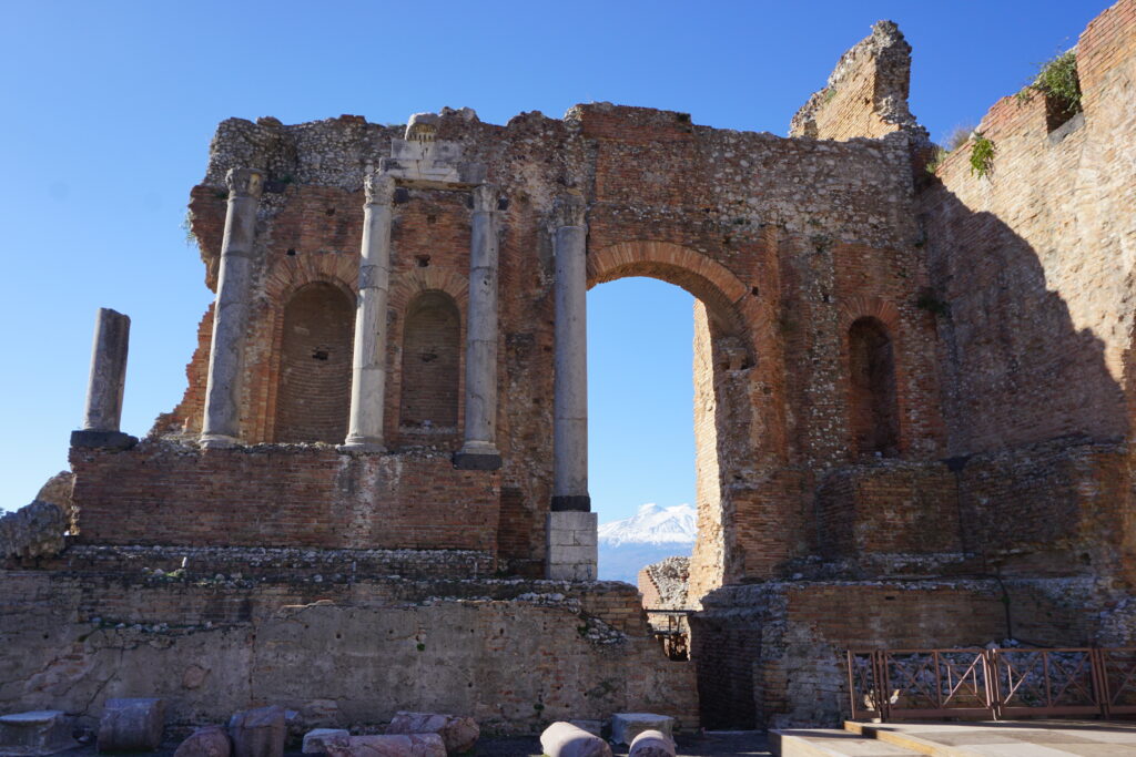 Ruins of the ancient Greek theatre of Taormina with its remaining tall, arched structures and columns against a clear blue sky, with the snowy peak of Mount Etna in the background.