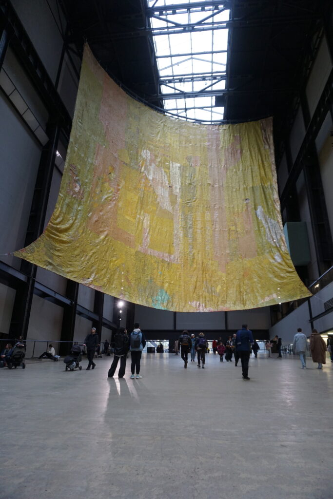 Visitors at the Tate Modern art gallery in London, observing a large, suspended, abstract textile art piece with a blend of golden, pink, and pale colors, under a spacious industrial ceiling with skylights.