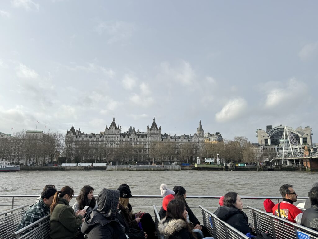 Passengers enjoying a river cruise on the Thames, with a view of the opulent riverside architecture and the London Eye in the distance, under a cloudy sky.