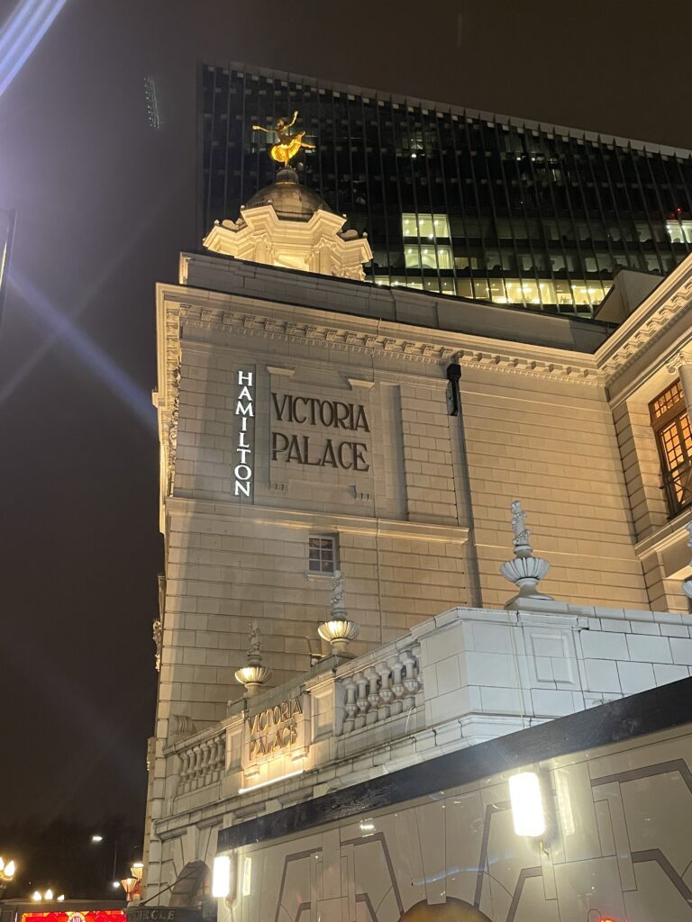 The illuminated facade of the Victoria Palace Theatre at night, showcasing its classic architecture with the name 'HAMILTON' prominently displayed, and the contrasting backdrop of a modern, high-rise building.