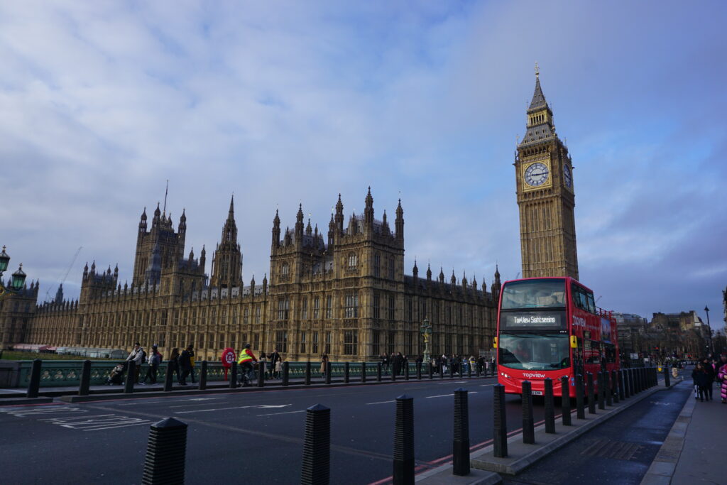 The iconic Palace of Westminster, with the Big Ben clock tower, under a cloudy sky as a red double-decker bus passes by on the street.