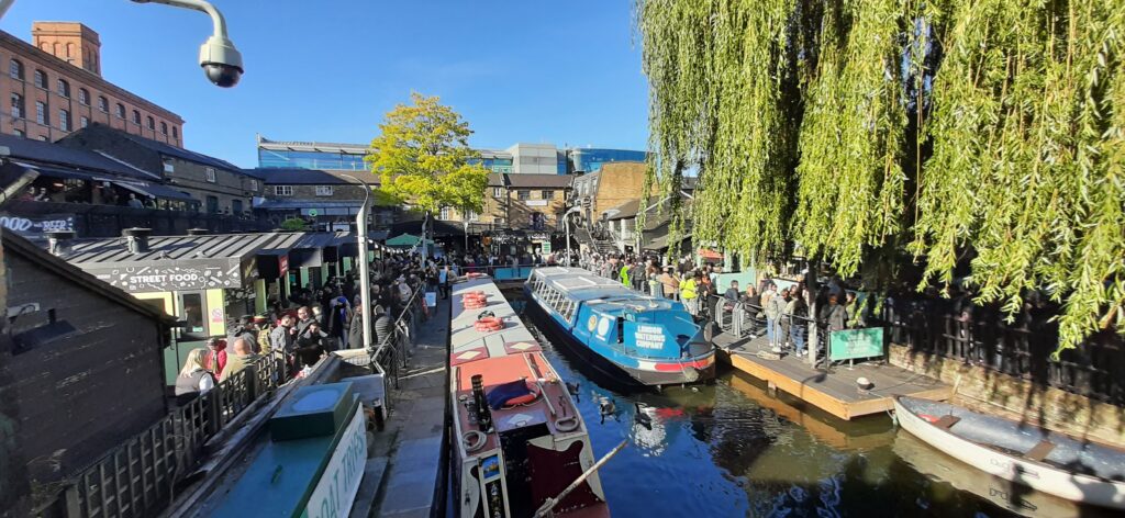 A panoramic view of Camden Market bustling with people, surrounded by colorful narrowboats moored along the Regent's Canal on a sunny day.