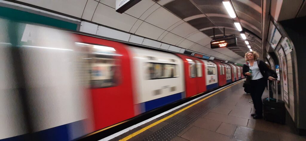 A motion-blurred image of a red London Underground train arriving at the station platform where a woman with a suitcase is waiting.