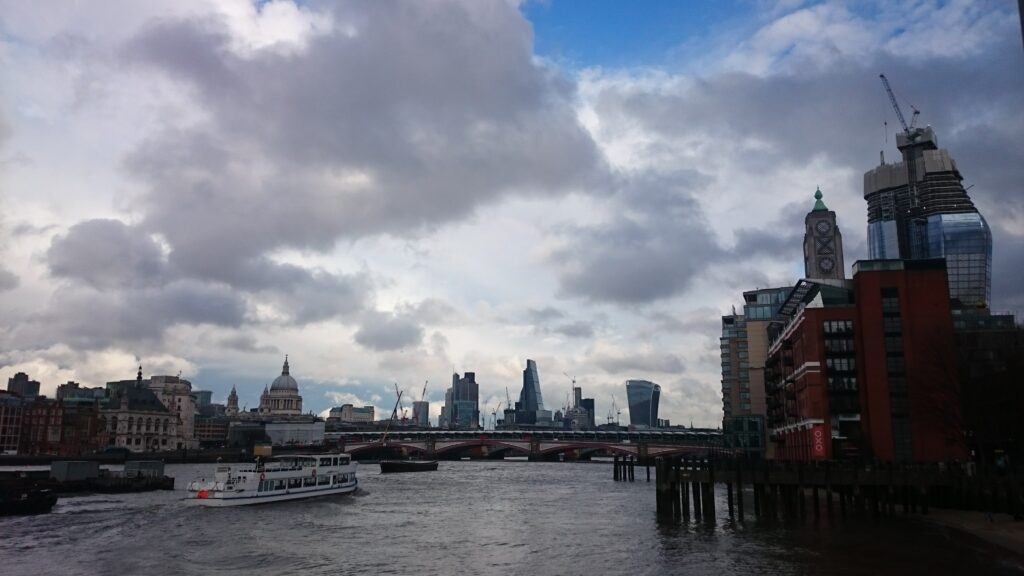 A cloudy sky over the River Thames, featuring a view of the London skyline with prominent buildings and a boat cruising on the water.