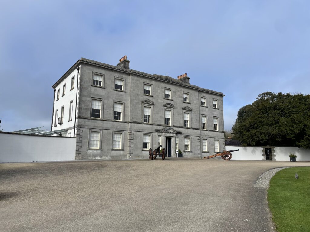 The exterior of the Battle of the Boyne Visitor center, A Historic grey stone mansion with a large front yard and a cannon display under a partly cloudy sky.