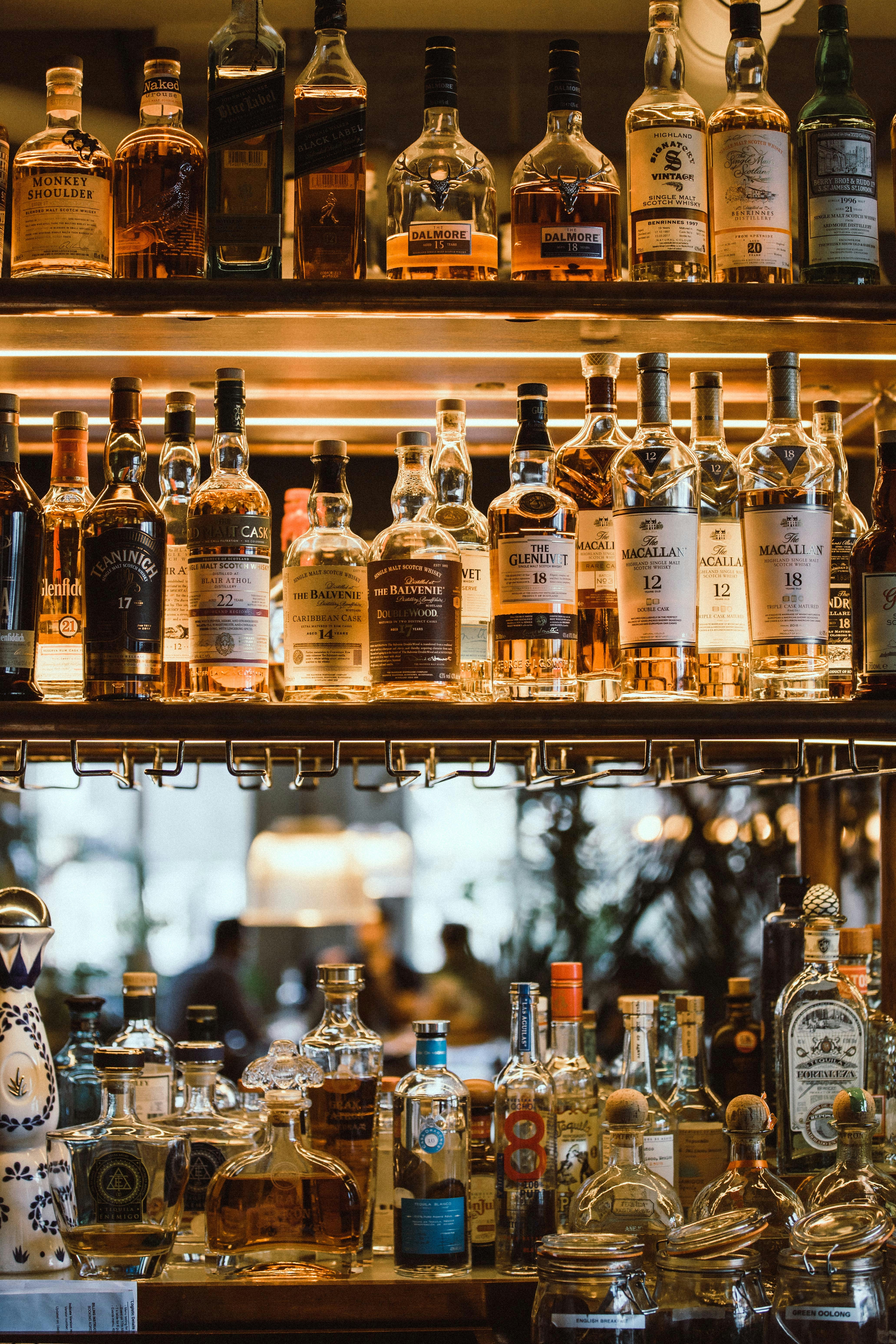 Elegant array of various whiskey and scotch bottles neatly displayed on backlit shelves above glass decanters, with the warm, ambient atmosphere of a bar in the background.