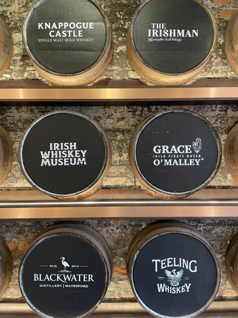 Shelves of labeled whiskey barrel ends showcasing various Irish whiskey distilleries, including Knappogue Castle and Blackwater, against a rustic brick wall.