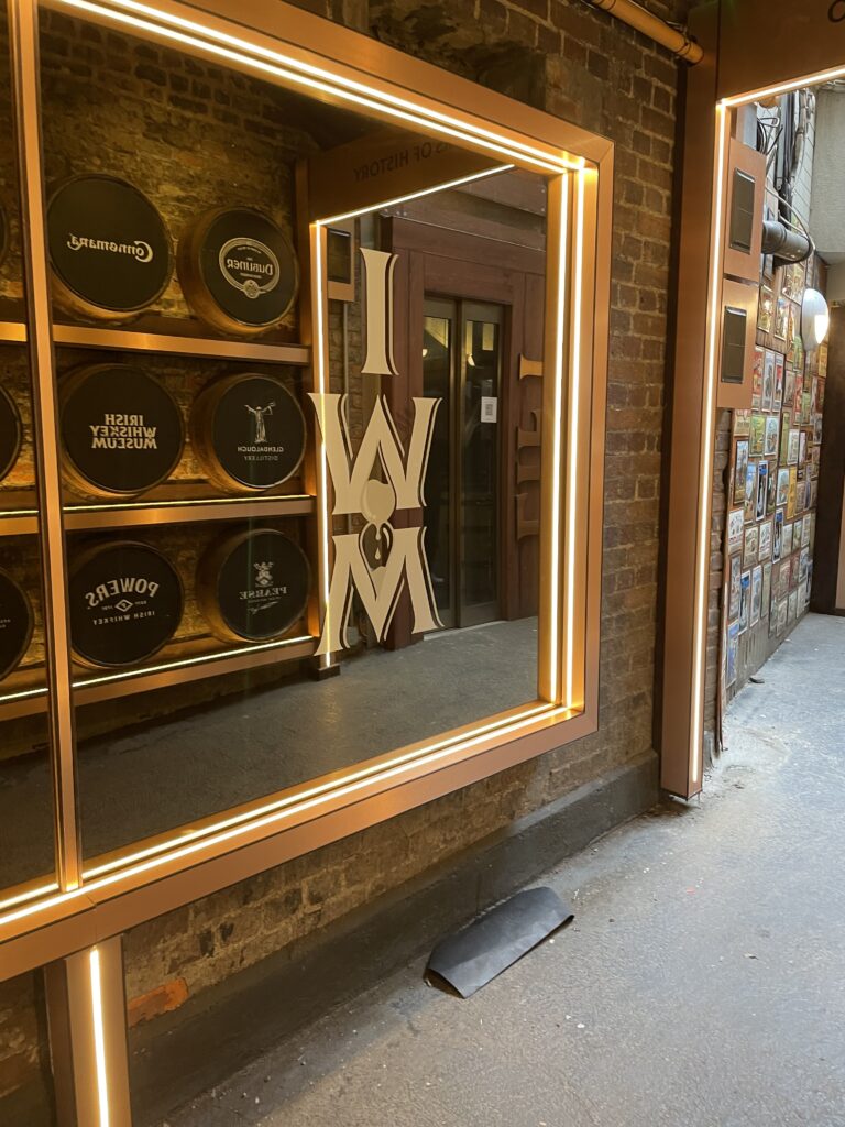 Entrance to the Irish Whiskey Museum with reflective gold lettering on the glass door, flanked by whiskey barrel ends on the walls, highlighting Irish distillery brands.