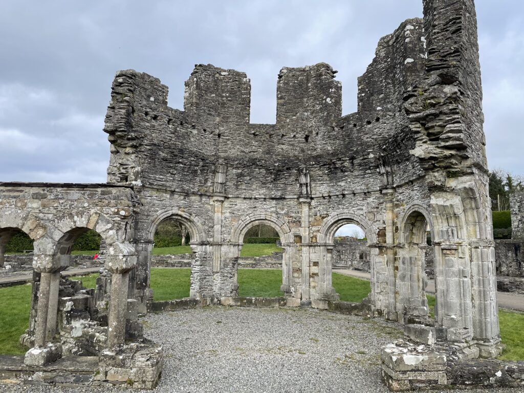 Ruins of Mellifont Abbey featuring remaining arches and columns with overgrown grass and a cloudy sky above.