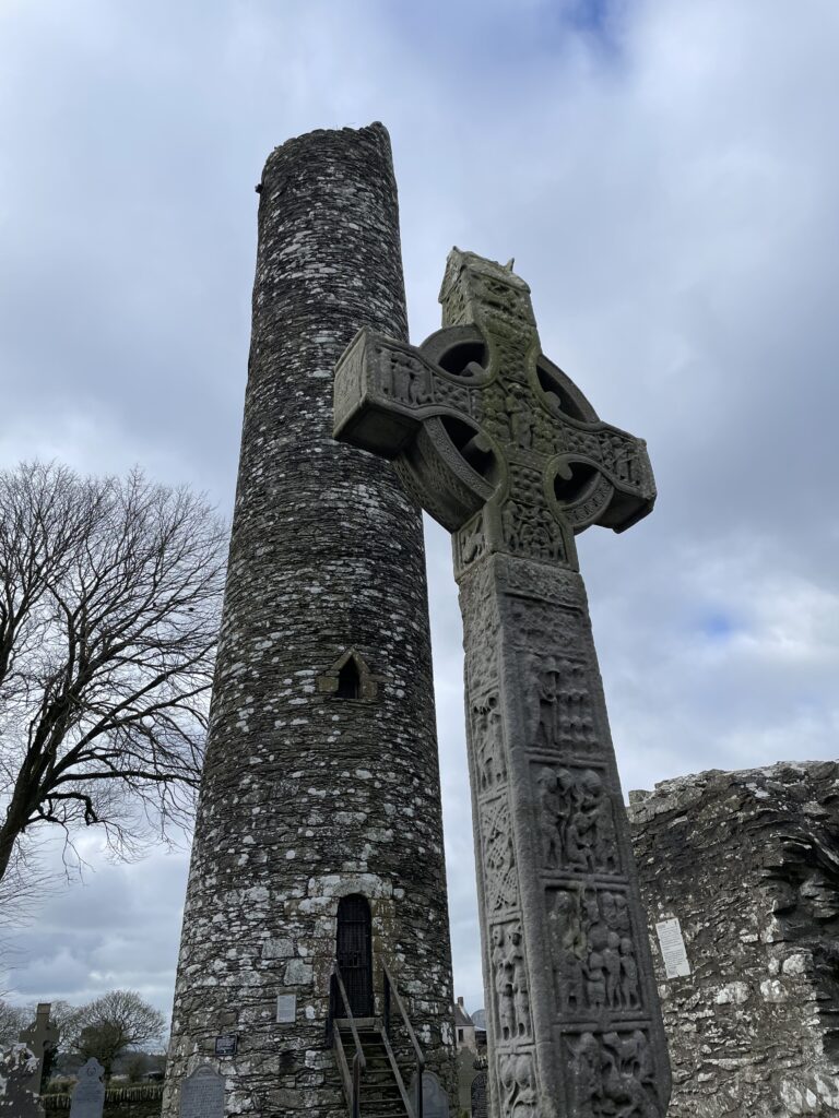 Ancient round tower of Monasterboice with a Celtic cross in the foreground against a cloudy sky.