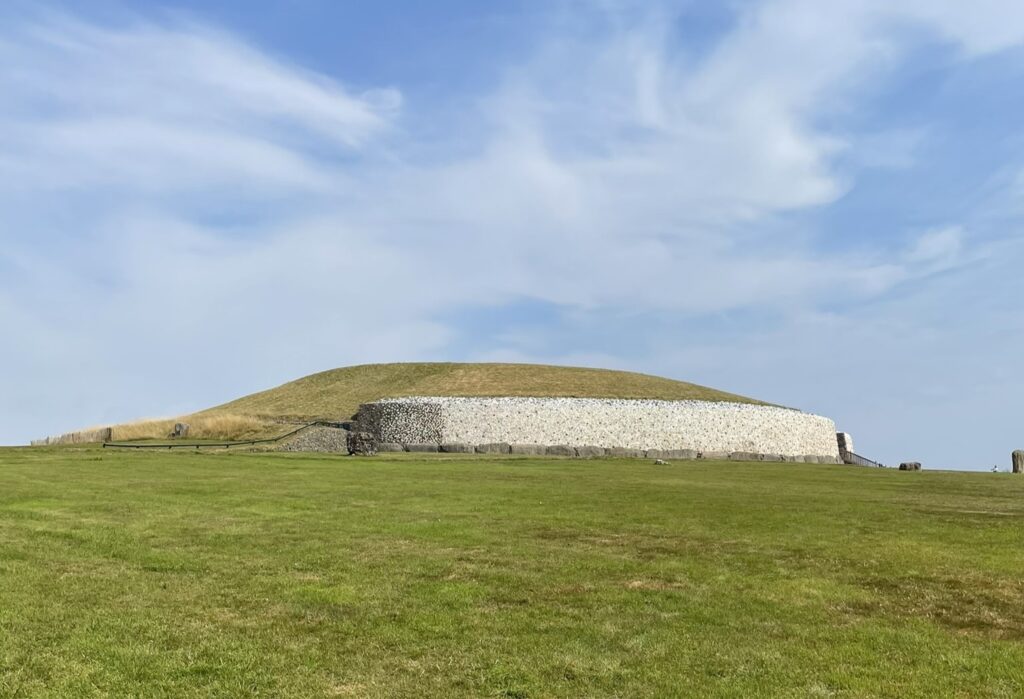 The ancient Newgrange neolithic mound in Ireland, with its iconic white quartz front wall, stands against a vibrant blue sky with scattered clouds, surrounded by a lush green field.