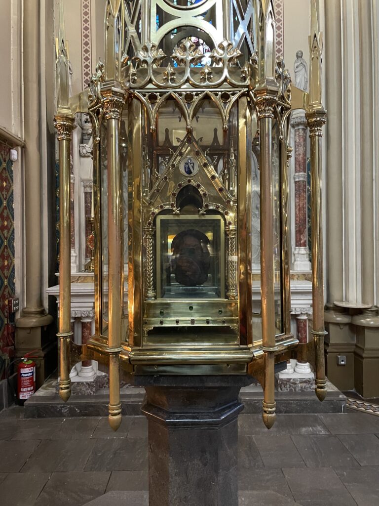 Ornate golden reliquary displayed on a pedestal inside a church with intricate designs and a reflection of stained glass windows.