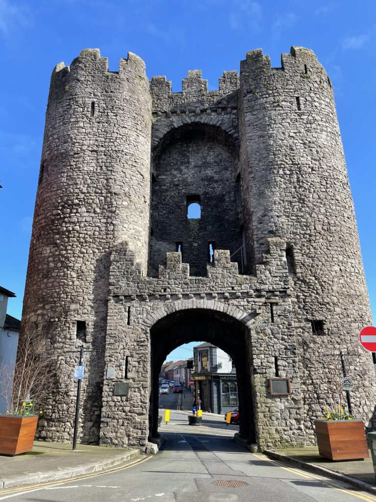 Medieval St. Laurence's Gate with twin turrets forming a stone archway over a street in bright daylight.