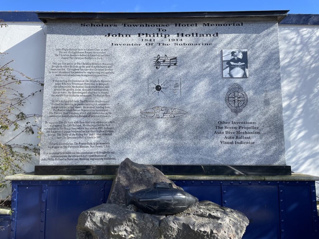 Memorial plaque for John Philip Holland, inventor of the submarine, featuring text, diagrams, and images on a wall with an emblem and propeller design.