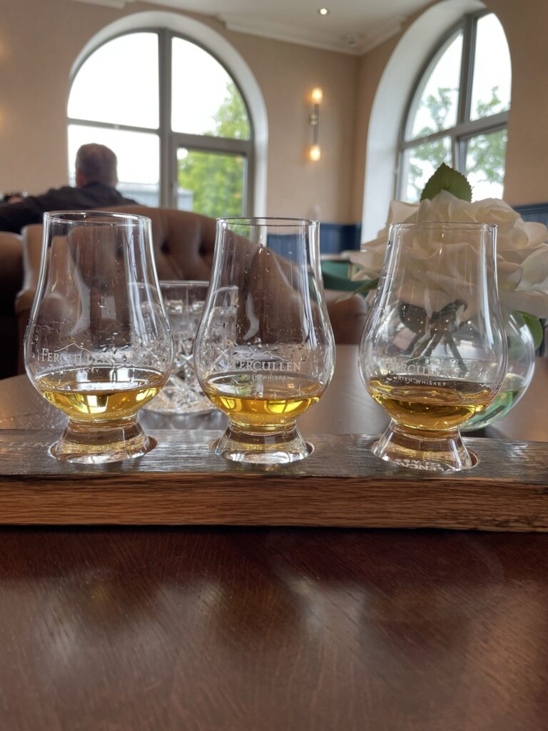 Three glasses of amber whiskey lined up on a wooden table, each labeled with a different distillery name, ready for tasting on the best whiskey tour in Dublin.