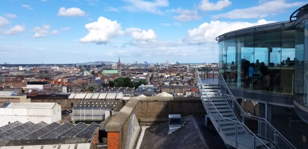 Panoramic view from the Guinness Storehouse in Dublin, showcasing a sweeping cityscape under a partly cloudy sky. To the right, visitors are seen through the glass walls of the observation deck, with the iconic metal staircases leading up to the viewing area.