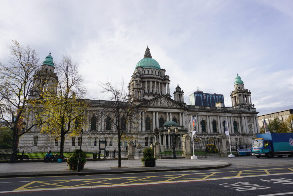 The Belfast City Hall stands majestic with its Baroque Revival architecture, featuring a prominent green dome and four corner towers, set against a backdrop of overcast sky. In the foreground, yellow leaves adorn sparse trees and vehicles pass by on the road, indicating a quiet day in the city.