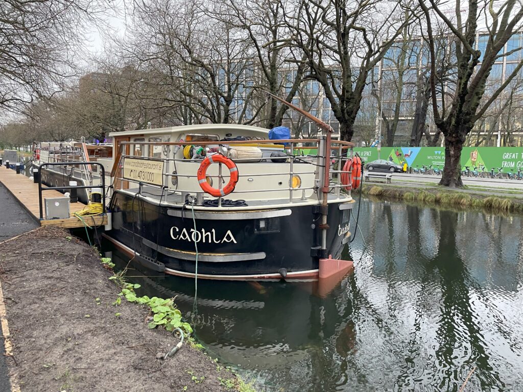 A vintage-styled canal boat named 'Capella' is moored on the tranquil waters of a tree-lined canal. In the background, modern buildings peek through the bare branches, and an advertisement is visible on a green fence, juxtaposing urban development with the leisurely pace of boat life.