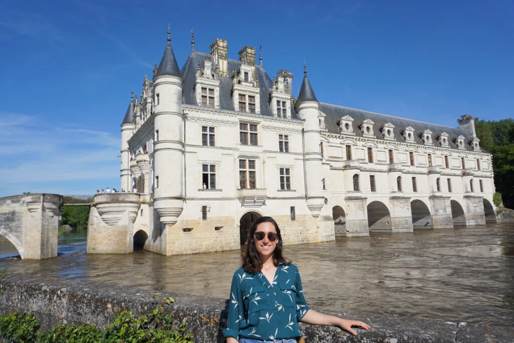 The author, wearing a green blouse, stands in front of a French castle built across a river. The scene suggests the importance of packing casual yet stylish clothing for Europe in spring, with layers to adapt to varying weather.