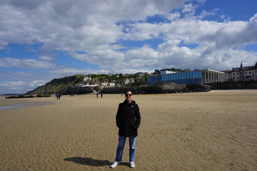 A woman in a black waterproof rain jacket, jeans, and white sneakers stands on the beach at Port Winston smiling at the camera. Behind her is a cloud filled sky with patches of blue poking through. The photo depicts the changeable weather found across Europe in spring and underscores the importance of packing layers for spring travel.