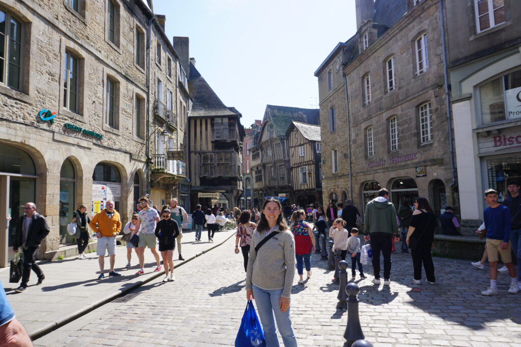 The author stands in the streets of St. Malo, France, surrounded by other pedestrians walking about. She is holding a blue tote bag filled with purchases from the local market. 