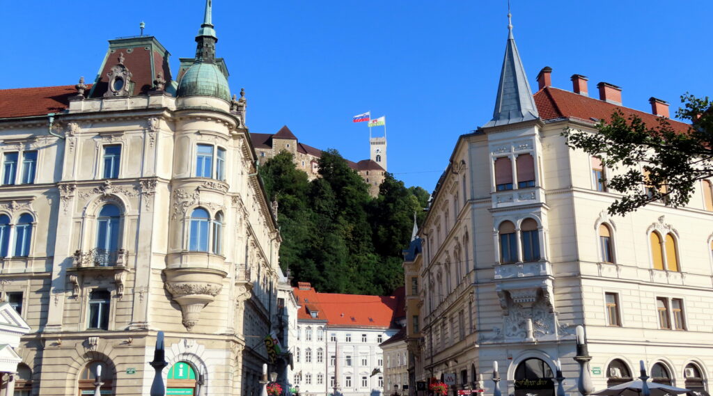 Ljubljana Castle Viewed From The Triple Bridge, The castle rises above on a tree-lines hill with two flags waving from a tower. In the foreground, beautiful buildings flank the hillside.