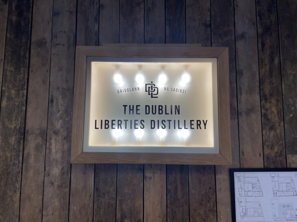 Illuminated sign on wooden wall reads "The Dublin Liberties Distillery" with decorative lights. Understated elegance and warmth. Architectural plans visible below.